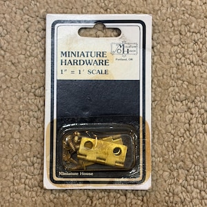 Puede incluir: Juego de herrajes en miniatura en un paquete de plástico transparente. El paquete tiene un borde blanco y negro e incluye el texto "MINIATURE HARDWARE 1" = 1' SCALE". El juego incluye pequeñas bisagras y tornillos de latón.