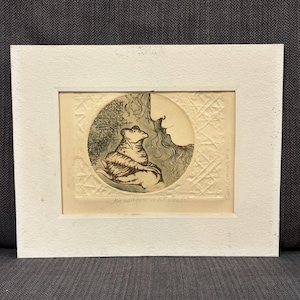 May include: A framed etching on cream paper featuring a circular illustration of a frog resting in a hand, with textured cross-hatched patterns in the background. The print includes the handwritten text My banquet is G.F. Handel and Carl Luminus 1970.