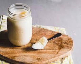 May include: A clear glass jar filled with a creamy, off-white substance sits on a wooden heart-shaped chopping board. A small dollop of the substance rests on a spoon beside the jar. The board rests on a yellow and white striped cloth.