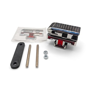 Puede incluir: Un emblema cromado y de fibra de carbono con detalles en rojo, junto con los accesorios de montaje. El emblema tiene la forma de una letra "T" estilizada. Se incluye un soporte negro, dos varillas roscadas y dos tuercas.