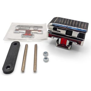 May include: A chrome and red GT emblem with a dark wood-grain top section, accompanied by a separate GT emblem on a clear backing, a black mounting bracket, two threaded metal rods, and two small silver hex nuts for vehicle installation.