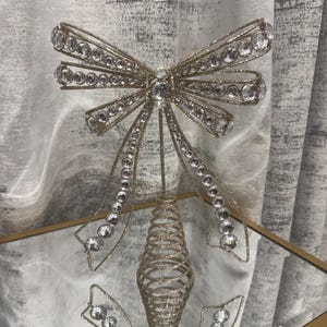 May include: A decorative gold-toned metal bow topper featuring a glittery finish and embedded clear circular crystals. The bow is mounted on a spiraled, cone-shaped base, designed to sit atop a surface with a reflective metallic structure.