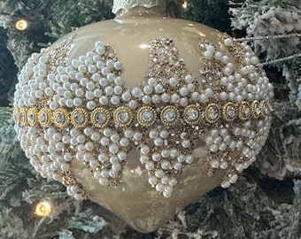 May include: A champagne-coloured glass Christmas ornament with a teardrop shape. The surface is decorated with a horizontal band of gold-toned metal featuring embedded clear rhinestones, surrounded by clusters of white faux pearls and gold glitter.