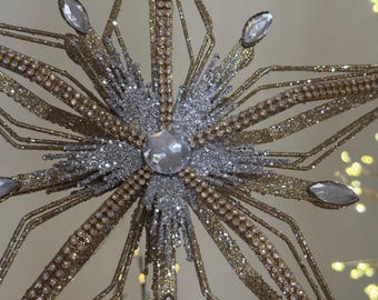 Puede incluir: Primer plano de un adorno decorativo en forma de estrella. La estrella presenta un acabado dorado brillante con detalles plateados y adornos transparentes en forma de gema. La estrella está diseñada con múltiples puntas y una gema central grande.