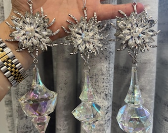 May include: Three decorative ornaments with silver chains. Each ornament features a sparkling, multi-faceted crystal design. The top of each ornament has a silver, starburst-shaped design. The bottom of each ornament has a clear, prism-like, teardrop shape.