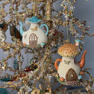 May include: Two whimsical mushroom-shaped teapot ornaments with intricate details, hanging on a sparkling, gold-toned tree. One has a blue top and the other an orange top. Both have butterfly accents and miniature doors.