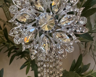 May include: A silver-toned decorative ornament featuring a large central faceted crystal surrounded by marquise-cut and round clear stones arranged in a floral pattern, with multiple long, dangling strands of small clear crystals hanging below.