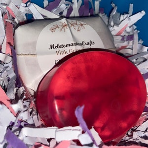 May include: A pink grapefruit scented soap bar in a clear wrapper, with a white label that reads "MeletomanianCrafts Pink Grapefruit Clay". The soap is sitting in pink and purple shredded paper.