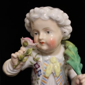 May include: A porcelain figurine of a child holding a pink rose. The child wears a white jacket with floral and striped details, a white shirt, and a purple skirt. The figurine has rosy cheeks and white curly hair.