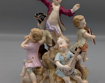 Op de afbeelding: Een porseleinen beeldje met vier kinderen die muziekinstrumenten bespelen op een rotsachtige basis. Eén kind in een bordeauxrode jas staat bovenaan, terwijl anderen in roze, blauwe en gedessineerde kleding fluit, mandoline en viool spelen.