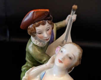 May include: A porcelain figurine depicting a man playing a lute and a woman with a rose in her hair. The man wears a green coat and a brown hat. The woman is in a white dress. The figurine has a black background.