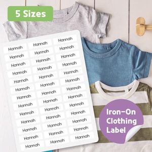 May include: A sheet of white iron-on clothing labels with the name "Hannah" printed on them, next to several children's t-shirts. The labels are rectangular and the text "Iron-On Clothing Label" is visible. The text "5 Sizes" is also visible.