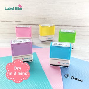 Könnte beinhalten: Eine Sammlung farbiger Namensstempel mit den Namen Abigail, Nicholas, Julian, Harper und Thomas. Die Stempel sind rosa, gelb, grün, lila und blau. Der Text "Dry in 3 mins" befindet sich in einer rosa Wolke.