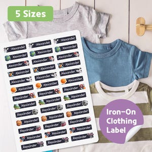 May include: A sheet of iron-on clothing labels with the name "Alexander" and space-themed graphics. The labels are black with white text and colorful illustrations of planets, rockets, and astronauts. The image also shows several children's t-shirts.