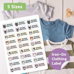 May include: Sheet of iron-on clothing labels with various cat illustrations and the name "Benjamin Armstrong." The labels are displayed with several children's t-shirts in different colors. Text on the labels reads "Iron-On Clothing Label" and "5 Sizes."