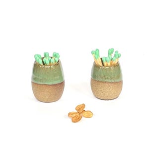 May include: Two small, rounded ceramic vessels with a two-tone glaze featuring a textured, sandy-brown base and a smooth, sage-green upper section. Each vessel is filled with wooden matches that have light green tips. A dried flower petal lies below.