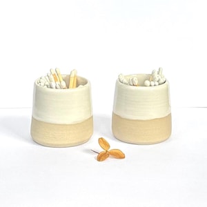 May include: Two small, cylindrical ceramic cups with a two-tone finish. The upper portion is glazed in a glossy off-white, while the lower base remains in a matte, unglazed beige clay. One cup holds wooden matches, the other holds cotton swabs.
