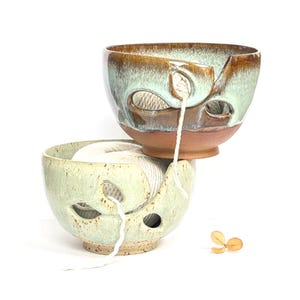 May include: Two ceramic yarn bowls, one stacked on the other. The top bowl is brown and green, the bottom bowl is green. Both have cut-out designs for yarn to feed through. White yarn is visible in the bowls.