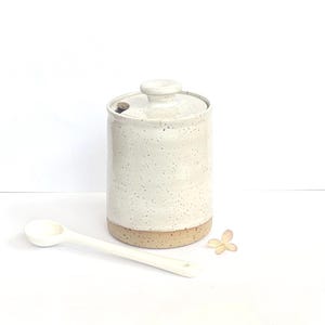 May include: A cylindrical ceramic jar with a speckled off-white glaze and a matching lid featuring a small knob handle and a side notch. The base is unglazed, revealing a tan speckled clay texture. A long, slender white ceramic spoon rests nearby.
