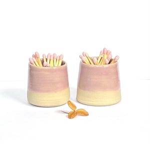 May include: Two small ceramic cups with a two-tone glaze featuring a soft pink upper section and a cream-coloured base. Each cup is filled with wooden matches that have light pink tips. A single dried brown leaf lies on the surface between them.