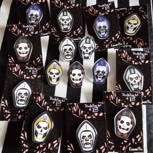 Könnte beinhalten: Eine Sammlung von Emaille-Anstecknadeln mit Totenkopf-Designs und unterschiedlich farbigen Mitren. Die Anstecknadeln werden auf schwarzen Karten mit einem rot-weißen Blumenmuster präsentiert. Text: "Doodle Bug Art of KIM" und "Enamel Pin".