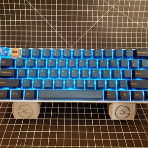 May include: A compact, backlit mechanical keyboard with blue keycaps and a white frame. The keys are illuminated with a bright blue light. The keyboard rests on two gray, decorative stands. The Tab key has a unique design.