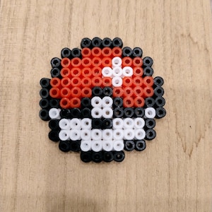 Op de afbeelding: Een handgemaakte, gepixelde Pokeball gemaakt van rode, zwarte, witte en oranje kralen. De kralenkunst heeft een wit kruis op de bovenste helft en een witte knop op de onderste helft, tegen een houten achtergrond.