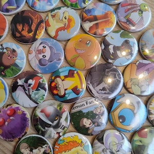 Op de afbeelding: Een verzameling ronde, kleurrijke buttonspins met verschillende stripfiguren. De buttons hebben een glanzende afwerking en zijn gerangschikt op een lichtbruin oppervlak. De ontwerpen omvatten verschillende wezens en personages.
