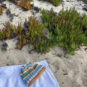 May include: A knit beanie featuring horizontal stripes in shades of blue, green, yellow, orange, and white. The hat has a ribbed cuff at the bottom and a textured, chunky yarn construction, resting on a plain white fabric surface.