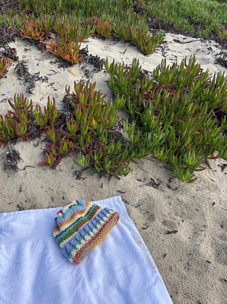 May include: A knit beanie featuring horizontal stripes in shades of blue, green, yellow, orange, and white. The hat has a ribbed cuff at the bottom and a textured, chunky yarn construction, resting on a plain white fabric surface.