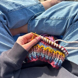 May include: A person is knitting a colorful striped scarf with a pair of gold knitting needles. The scarf is made with a variety of yarns in shades of pink, blue, yellow, and white.