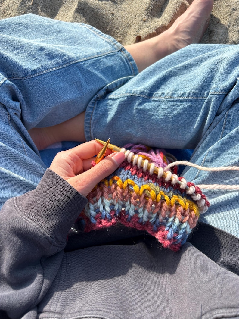 May include: A person is knitting a colorful striped scarf with a pair of gold knitting needles. The scarf is made with a variety of yarns in shades of pink, blue, yellow, and white.