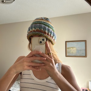 May include: A chunky knit beanie featuring a multi-colored striped pattern in shades of blue, cream, yellow, and rust orange. The hat has a thick, ribbed folded brim and a textured, hand-knitted appearance with a soft, cozy look and feel.
