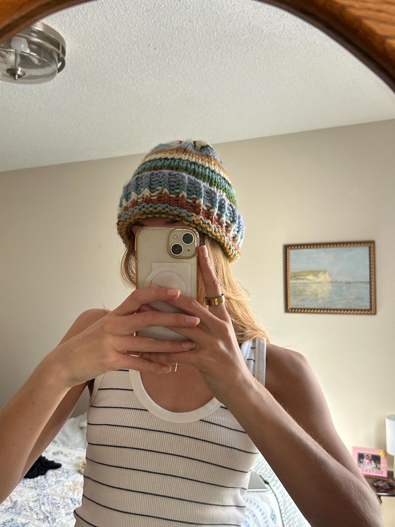 May include: A chunky knit beanie featuring a multi-colored striped pattern in shades of blue, cream, yellow, and rust orange. The hat has a thick, ribbed folded brim and a textured, hand-knitted appearance with a soft, cozy look and feel.
