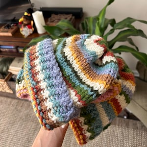 May include: A thick, hand-crocheted beanie featuring horizontal stripes in a palette of cream, light blue, forest green, mustard yellow, and terracotta orange. The knit texture is prominent, with a folded brim and a rounded crown construction.