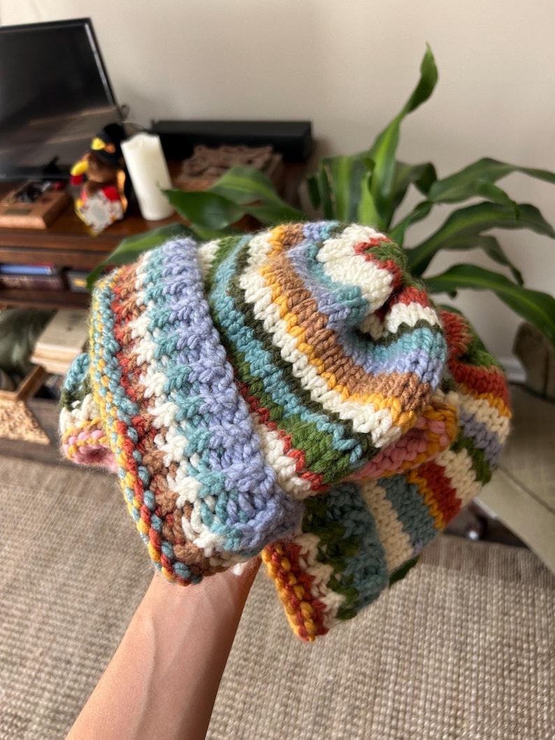 May include: A thick, hand-crocheted beanie featuring horizontal stripes in a palette of cream, light blue, forest green, mustard yellow, and terracotta orange. The knit texture is prominent, with a folded brim and a rounded crown construction.