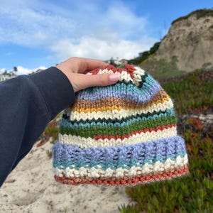 May include: A hand-knitted beanie featuring horizontal stripes in various colors including light blue, cream, mustard yellow, forest green, and rust orange. The hat has a thick, folded ribbed brim and a chunky knit texture throughout the entire piece.