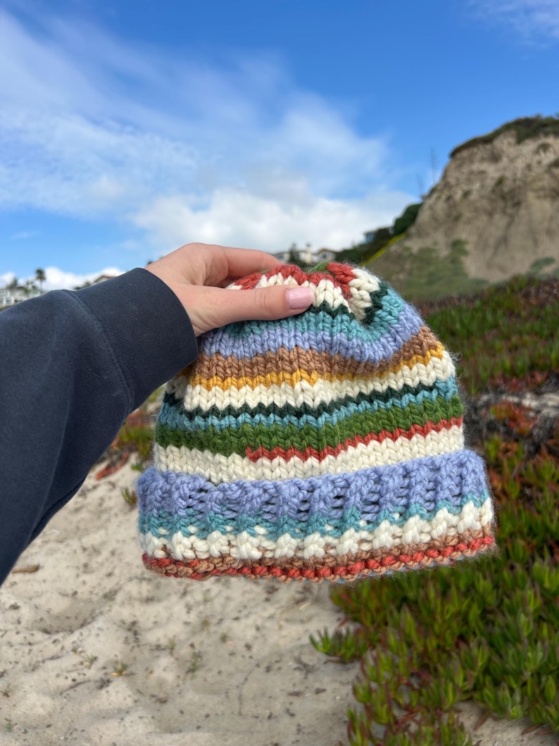 May include: A hand-knitted beanie featuring horizontal stripes in various colors including light blue, cream, mustard yellow, forest green, and rust orange. The hat has a thick, folded ribbed brim and a chunky knit texture throughout the entire piece.