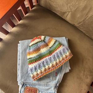 May include: A multi-colored striped knit beanie with a folded brim featuring horizontal bands of lavender, green, orange, and cream yarn, resting on top of a pair of light blue denim jeans with a visible brown leather brand patch on the back pocket.