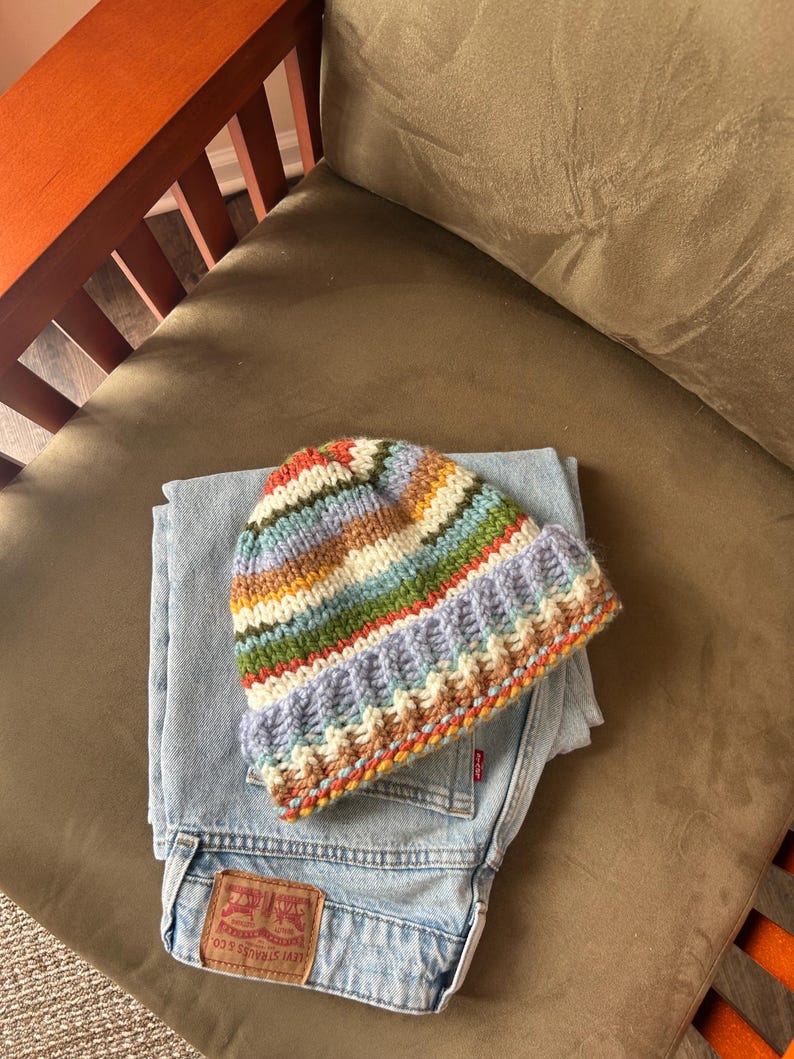 May include: A multi-colored striped knit beanie with a folded brim featuring horizontal bands of lavender, green, orange, and cream yarn, resting on top of a pair of light blue denim jeans with a visible brown leather brand patch on the back pocket.