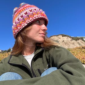 May include: A person wearing a colorful knitted beanie hat and a green fleece jacket. The person is sitting with their legs crossed and looking off to the side.