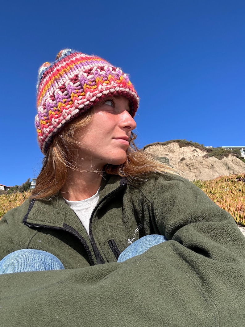 May include: A person wearing a colorful knitted beanie hat and a green fleece jacket. The person is sitting with their legs crossed and looking off to the side.