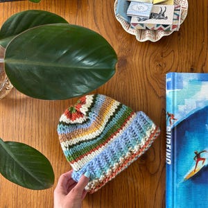 May include: A hand-knitted beanie featuring horizontal stripes in shades of blue, green, yellow, white, and rust orange. The hat has a thick, ribbed brim and a textured knit pattern throughout, showcasing a multi-colored yarn design.