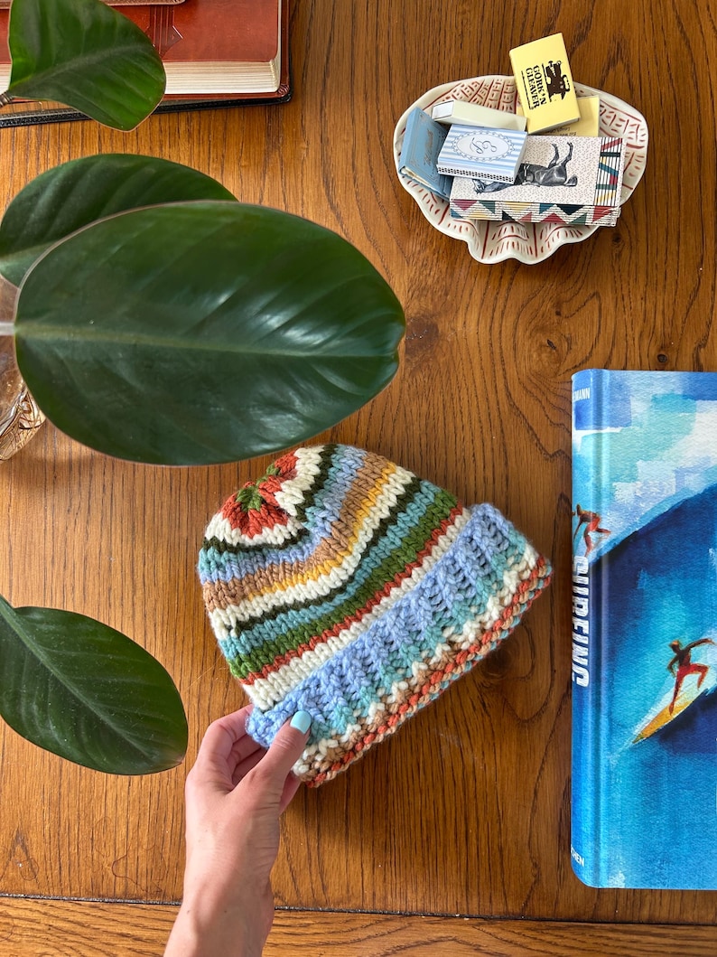 May include: A hand-knitted beanie featuring horizontal stripes in shades of blue, green, yellow, white, and rust orange. The hat has a thick, ribbed brim and a textured knit pattern throughout, showcasing a multi-colored yarn design.