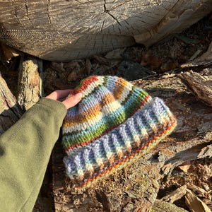 May include: A hand-knitted beanie featuring a multi-colored striped pattern in shades of blue, green, yellow, orange, and white. The hat has a thick, ribbed folded cuff and a textured knit construction made from chunky yarn in a variegated color scheme.