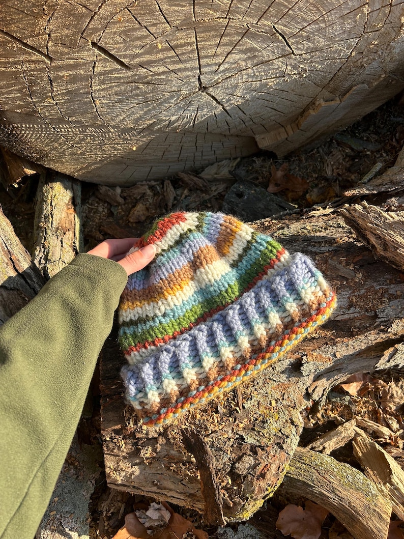 May include: A hand-knitted beanie featuring a multi-colored striped pattern in shades of blue, green, yellow, orange, and white. The hat has a thick, ribbed folded cuff and a textured knit construction made from chunky yarn in a variegated color scheme.