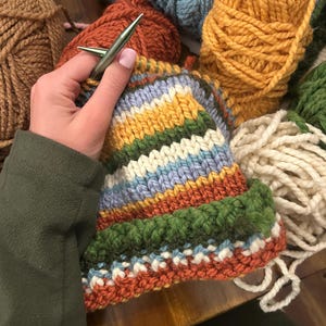 May include: A partially knitted hat featuring horizontal stripes in mustard yellow, forest green, cream, light blue, and burnt orange. The project is worked with metallic knitting needles, surrounded by several skeins of thick yarn in matching colors.