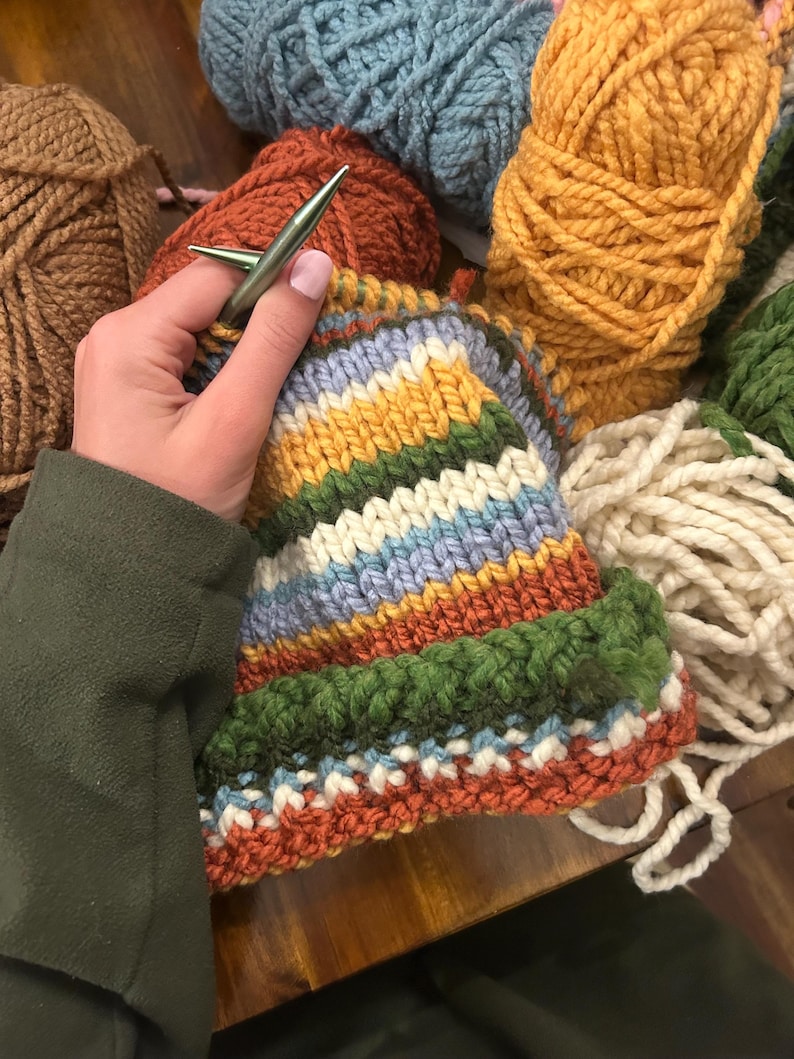 May include: A partially knitted hat featuring horizontal stripes in mustard yellow, forest green, cream, light blue, and burnt orange. The project is worked with metallic knitting needles, surrounded by several skeins of thick yarn in matching colors.