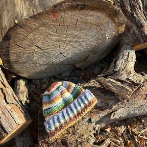 May include: A hand-knitted beanie featuring a variegated yarn pattern with horizontal stripes in shades of blue, green, yellow, orange, and white. The hat has a thick, ribbed cuff at the bottom and a rounded crown, crafted with a chunky knit texture.