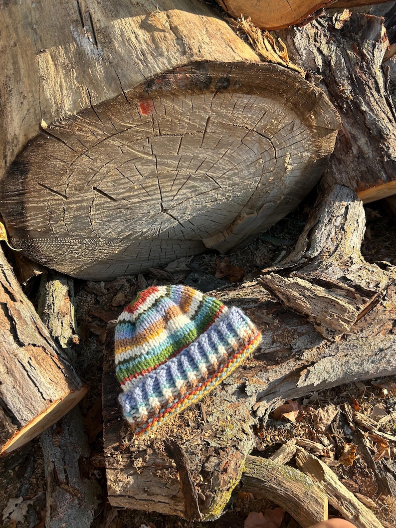 May include: A hand-knitted beanie featuring a variegated yarn pattern with horizontal stripes in shades of blue, green, yellow, orange, and white. The hat has a thick, ribbed cuff at the bottom and a rounded crown, crafted with a chunky knit texture.